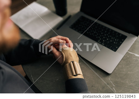 Close-up top view of male hands wearing wrist brace, massaging painful wrist while working remotely from home office, surrounded by laptop and notebook. Concept of struggles of remote work Close-up top view of male hands wearing wrist brace, massaging painful wrist while working remotely from home office, surrounded by laptop and notebook. Concept of struggles of remote work 125119843