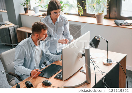 Man is sitting, woman is standing. Using pc. Two young designers is working together in the office Man is sitting, woman is standing. Using pc. Two young designers is working together in the office 125119861