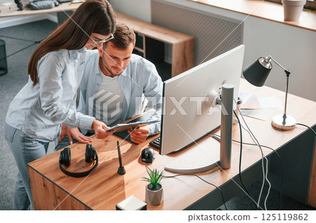 Man is sitting, woman is standing. Using pc. Two young designers is working together in the office 125119862
