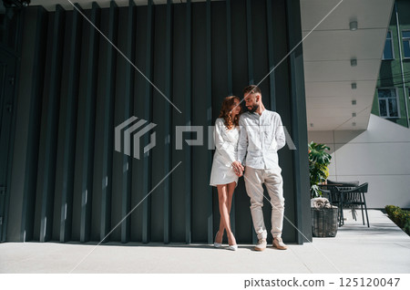 Near the modern building. Beautiful young couple is standing together against dark green background Near the modern building. Beautiful young couple is standing together against dark green background 125120047