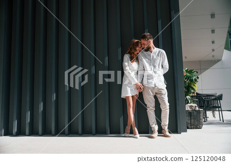 Near the modern building. Beautiful young couple is standing together against dark green background Near the modern building. Beautiful young couple is standing together against dark green background 125120048