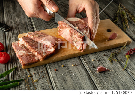 A person is skillfully cutting through gourmet meat on a wooden board, surrounded by fresh ingredients and spices, in a cozy kitchen environment during the evening 125120079