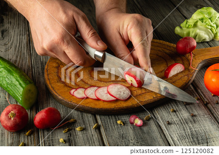 Hands skillfully slice vibrant radishes on a rustic wooden cutting board surrounded by an array of fresh vegetables including cucumbers and lettuce. Sunlight illuminates the work area Hands skillfully slice vibrant radishes on a rustic wooden cutting board surrounded by an array of fresh vegetables including cucumbers and lettuce. Sunlight illuminates the work area 125120082