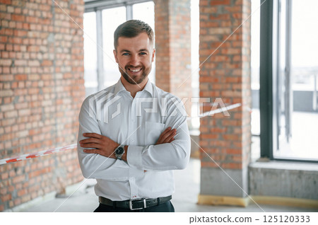 Standing with arms crossed. Man in white shirt is working indoors at construction site. Managing the project 125120333