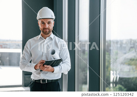 Standing near panoramic windows. Man in white shirt is working indoors at construction site. Managing the project Standing near panoramic windows. Man in white shirt is working indoors at construction site. Managing the project 125120342