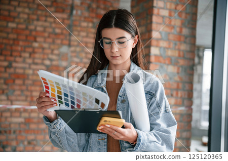 Notepad in hands. Young woman is standing in the unfinished building on construction site and working on a project Notepad in hands. Young woman is standing in the unfinished building on construction site and working on a project 125120365