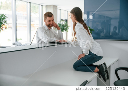 Woman sitting on the table on knees. Funny moment. Man in white shirt is ordering a room in the hotel by assistant help 125120443