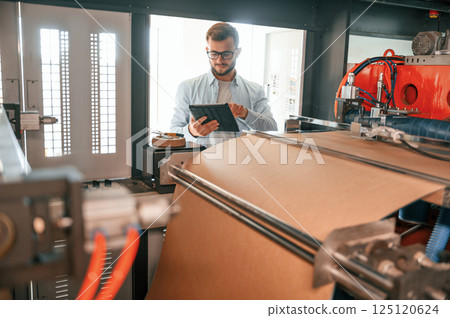 Holding digital tablet when standing near sheet cutting machine. Print house worker in white clothes is indoors Holding digital tablet when standing near sheet cutting machine. Print house worker in white clothes is indoors 125120624