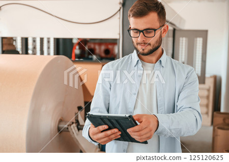 Holding digital tablet when standing near sheet cutting machine. Print house worker in white clothes is indoors 125120625