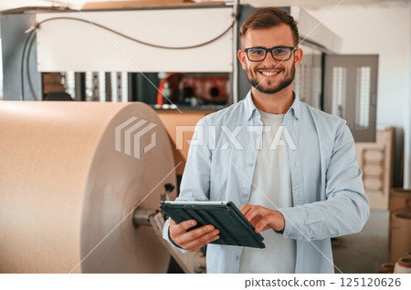 Holding digital tablet when standing near sheet cutting machine. Print house worker in white clothes is indoors 125120626
