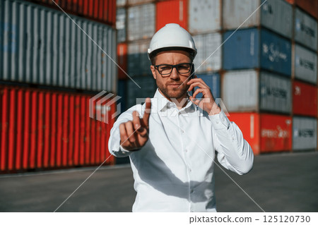 Showing a point finger while talking by phone. Male worker is on the location with containers 125120730