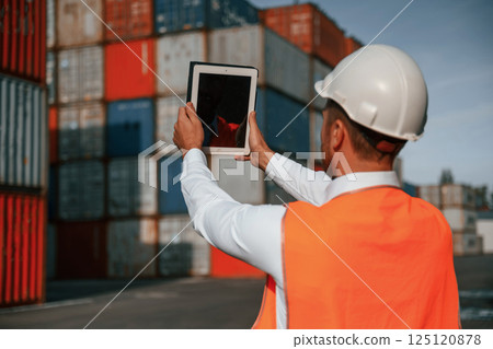 Holding tablet and making photo of the objects. Male worker is on the location with containers Holding tablet and making photo of the objects. Male worker is on the location with containers 125120878