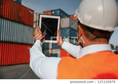 Holding tablet and making photo of the objects. Male worker is on the location with containers Holding tablet and making photo of the objects. Male worker is on the location with containers 125120879