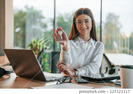 Holding keys in hands. Beautiful young woman in white clothes is working in office 125121117