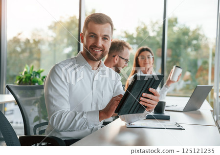 Man is looking at the camera. Three people in formal clothes are working in the modern office together 125121235