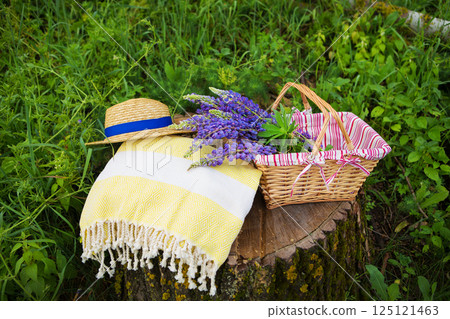 A summer picnic scene with a straw hat, yellow picnic blanket, wicker basket filled with lupines, set on a tree stump 125121463