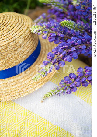 A straw hat with a blue ribbon rests on a yellow and white patterned blanket, adorned with vibrant purple lupine flowers A straw hat with a blue ribbon rests on a yellow and white patterned blanket, adorned with vibrant purple lupine flowers 125121464