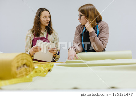 Two female seamstresses and fashion designers stand at table with roll of fabric negotiate Two female seamstresses and fashion designers stand at table with roll of fabric negotiate 125121477