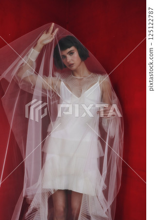 Elegant model poses in sheer fabric dress against a vibrant red background during a fashion photoshoot 125122787