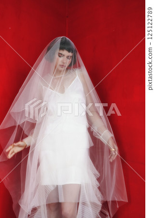 Bride in a white dress and sheer veil poses gracefully against a vibrant red backdrop 125122789