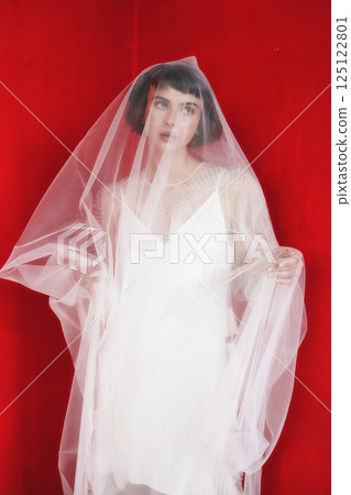 Elegant model in sheer veil against vibrant red backdrop during fashion photoshoot 125122801