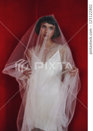 Model poses with delicate veil against a vibrant red backdrop showcasing elegance and contrast in fashion photography Model poses with delicate veil against a vibrant red backdrop showcasing elegance and contrast in fashion photography 125122802