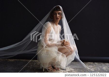 Stunning model in sheer white attire with veil posing gracefully on dark background in fashion studio 125122822