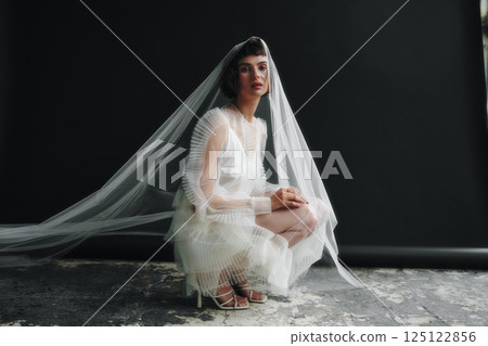 Model wearing a flowing white dress and veil poses in an urban studio during a fashion shoot in natural light Model wearing a flowing white dress and veil poses in an urban studio during a fashion shoot in natural light 125122856