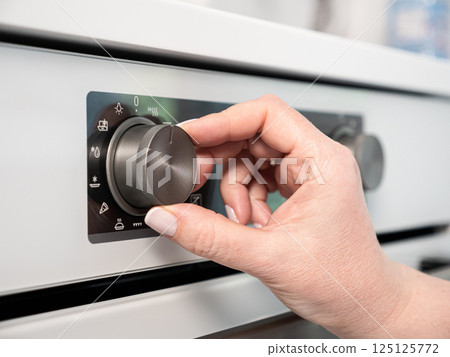 Woman adjusting modern white oven in kitchen.  Female hand pushing and turning knobs on oven control panel 125125772