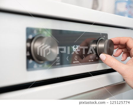 Woman adjusting modern white oven in kitchen.  Female hand pushing and turning knobs on oven control panel 125125774