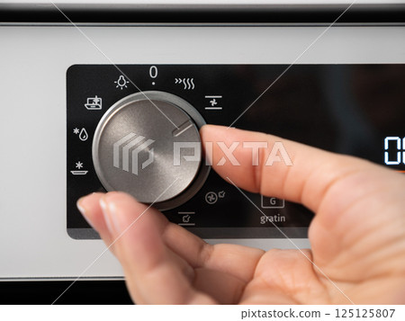 Woman adjusting modern white oven in kitchen.  Female hand pushing and turning knobs on oven control panel 125125807