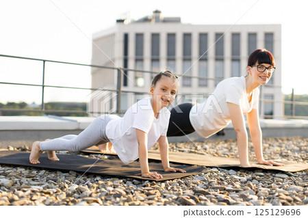 Mother and daughter performing exercise together on outdoor rooftop focusing on strength. Demonstrating healthy lifestyle and bonding during sports activity. Mother and daughter performing exercise together on outdoor rooftop focusing on strength. Demonstrating healthy lifestyle and bonding during sports activity. 125129696