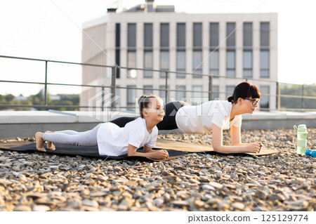 Mother and daughter doing plank exercises together outdoors on rooftop. Sunrise creates warm summer atmosphere. Women bonding through fitness and mindfulness, emphasizing health, family connection 125129774