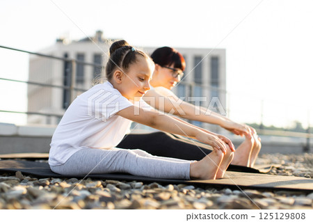 Mother and young daughter practicing stretching exercises on rooftop during sunny day. Family bonding moment promoting health, relaxation, and active lifestyle. 125129808