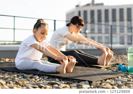 Adult woman and young girl stretching outdoors on yoga mats, promoting family fitness 125129809