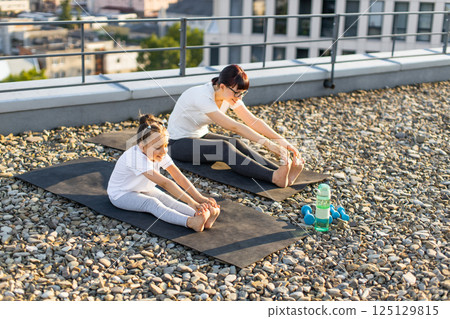 Mother and daughter practicing stretching exercises outdoors on rooftop during sunny day. Fitness mat and water bottle showing workout essentials. Mother and daughter practicing stretching exercises outdoors on rooftop during sunny day. Fitness mat and water bottle showing workout essentials. 125129815