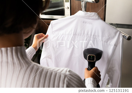 Close up view of woman's hand with steamer and summer white shirt, wife cleaning husband's clothes before he goes to work 125131191