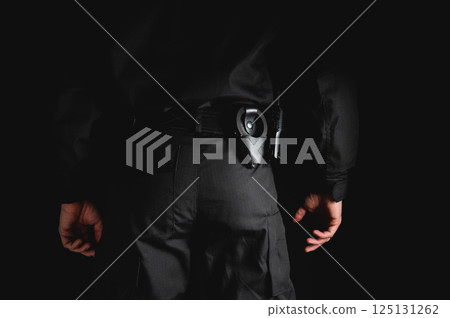 Police officer with handcuffs, a walkie-talkie and pepper spray on his belt stands against a black background. Close-up of a security guard ready to work Police officer with handcuffs, a walkie-talkie and pepper spray on his belt stands against a black background. Close-up of a security guard ready to work 125131262
