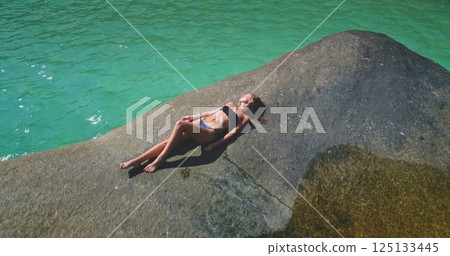 A person is reclining on a large rock, enjoying the warm sunlight by the stunning turquoise ocean. The tranquil setting creates a perfect atmosphere for relaxation. 125133445