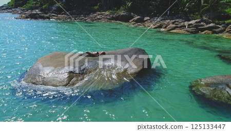 A person lies comfortably on a large rock, soaking up the sun undisturbed, surrounded by crystal-clear turquoise water at a serene tropical beach. Lush greenery adds to the picturesque backdrop. A person lies comfortably on a large rock, soaking up the sun undisturbed, surrounded by crystal-clear turquoise water at a serene tropical beach. Lush greenery adds to the picturesque backdrop. 125133447