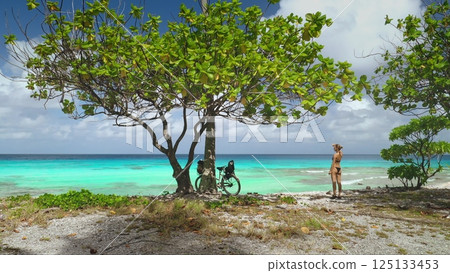 Captivating scene of a woman embracing the tranquility of a tropical paradise on rangiroa island, with her bicycle resting under the shade of a lush tree 125133453