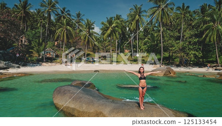 A woman stands on a rock, arms outstretched, at a picturesque beach surrounded by palm trees and clear turquoise water. It's a sunny day, perfect for relaxation and fun. 125133454