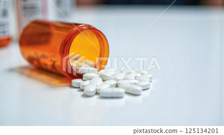 White tablets spilled from orange prescription bottle on clean surface in medical setting, concept of medication, healthcare, treatment, pharmacy, drug abuse prevention 125134201