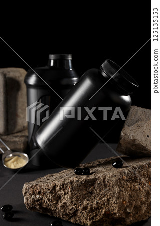 Black plastic jars, measuring spoon and pills on stones on black background 125135153