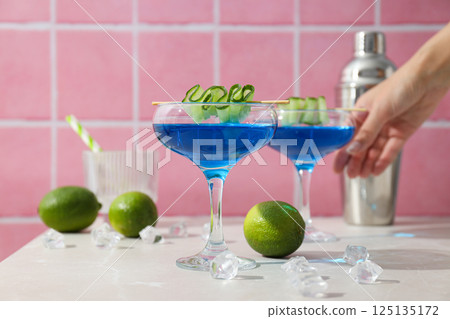 Blue cocktail with cucumber slice in glasses, hand, shaker, limes and ice cubes on pink background 125135172