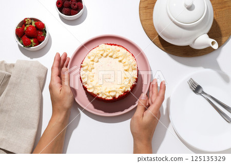 Red velvet cake on plate in female hands, bowls with berries and teapot on white background, top view Red velvet cake on plate in female hands, bowls with berries and teapot on white background, top view 125135329