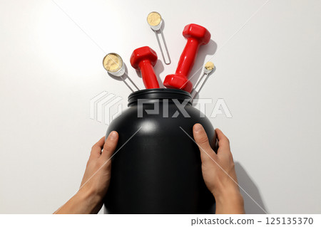 Black plastic jar in hands, dumbbells and measuring spoons on white background, top view Black plastic jar in hands, dumbbells and measuring spoons on white background, top view 125135370