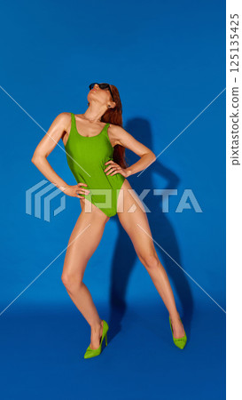Full-length photo of young beautiful woman wearing in green swimsuit and heels stretching back in dramatic pose on blue background. Full-length photo of young beautiful woman wearing in green swimsuit and heels stretching back in dramatic pose on blue background. 125135425