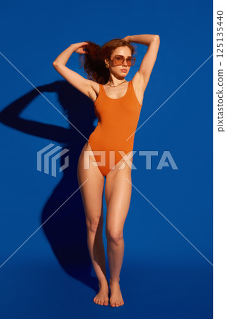 Graceful woman with confident stance posing barefoot in orange swimsuit with strong sunlight and motion against blue studio background. 125135440