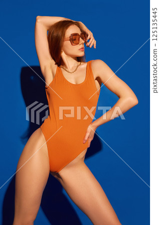 Red-haired woman wearing orange swimsuit and chunky sunglasses leaning back with hand on hip and body glowing against blue studio background. 125135445